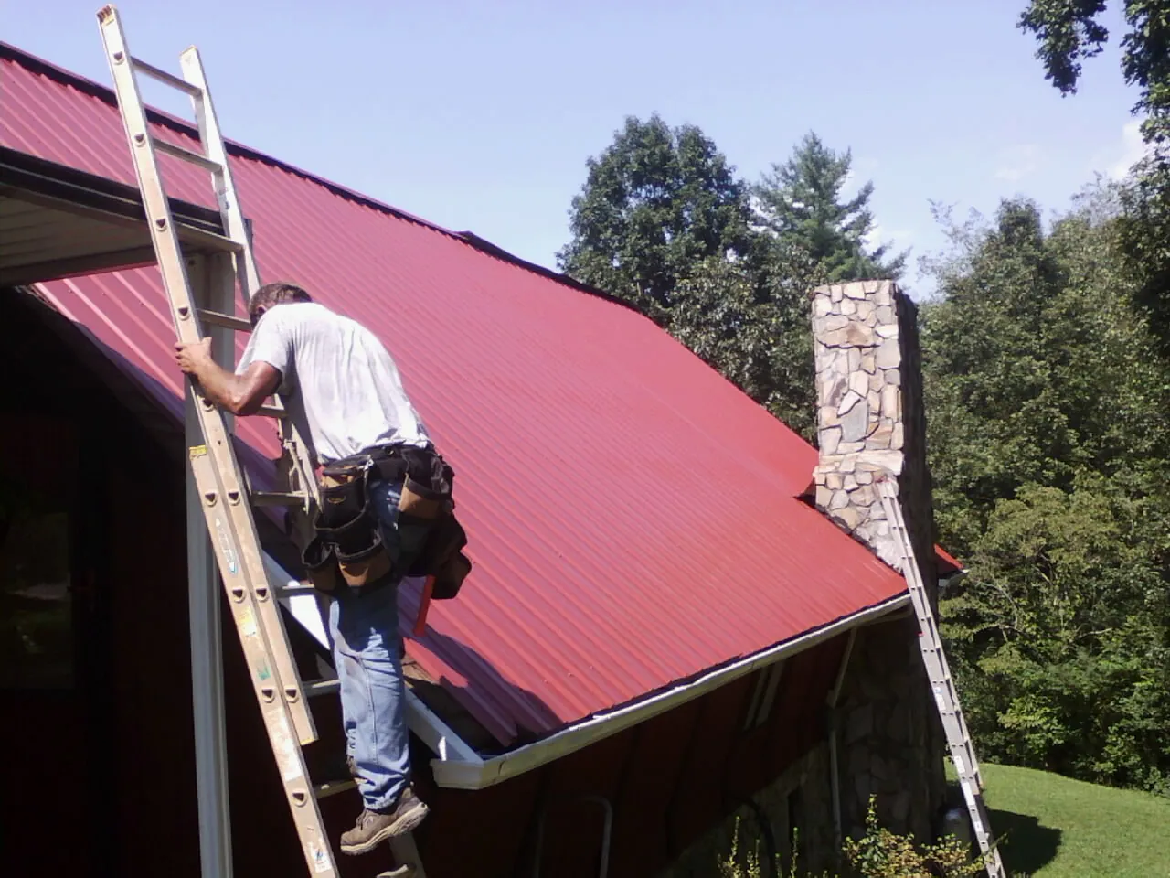 Roofer on a ladder beside a red metal roof and stone chimney — Soffit & Fascia Repair by Roofing Brothers in Gwynedd